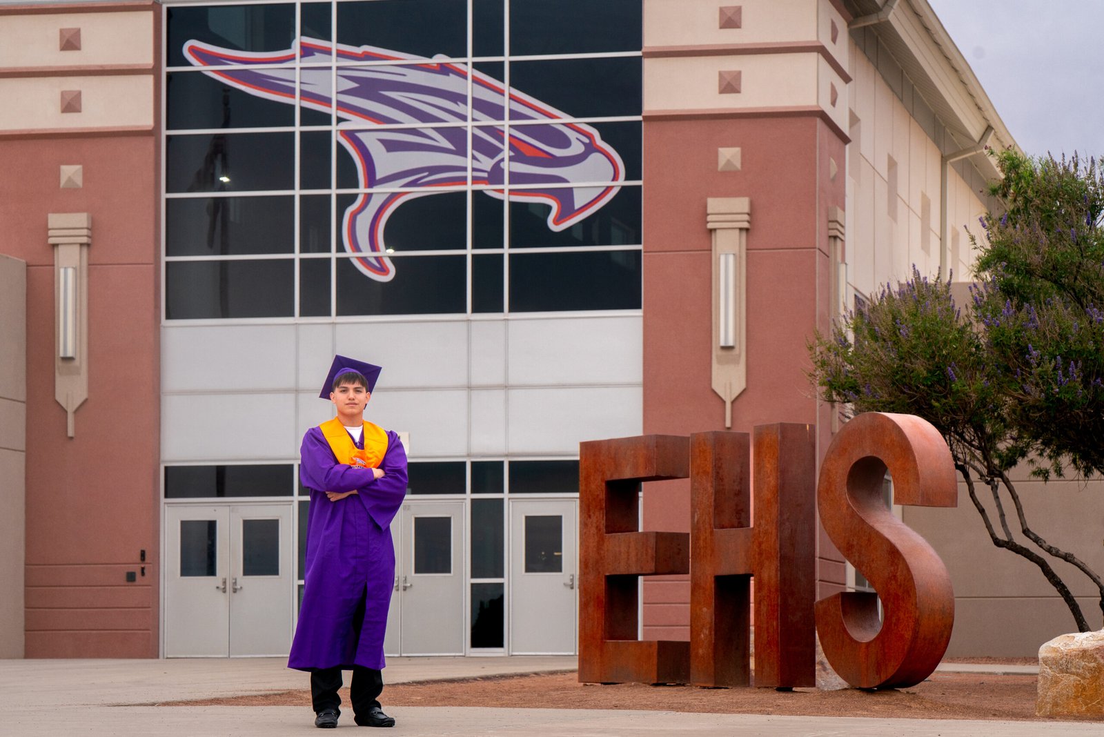 Graduate in purple cap and gown standing beside EHS lettering and an eagle emblem.