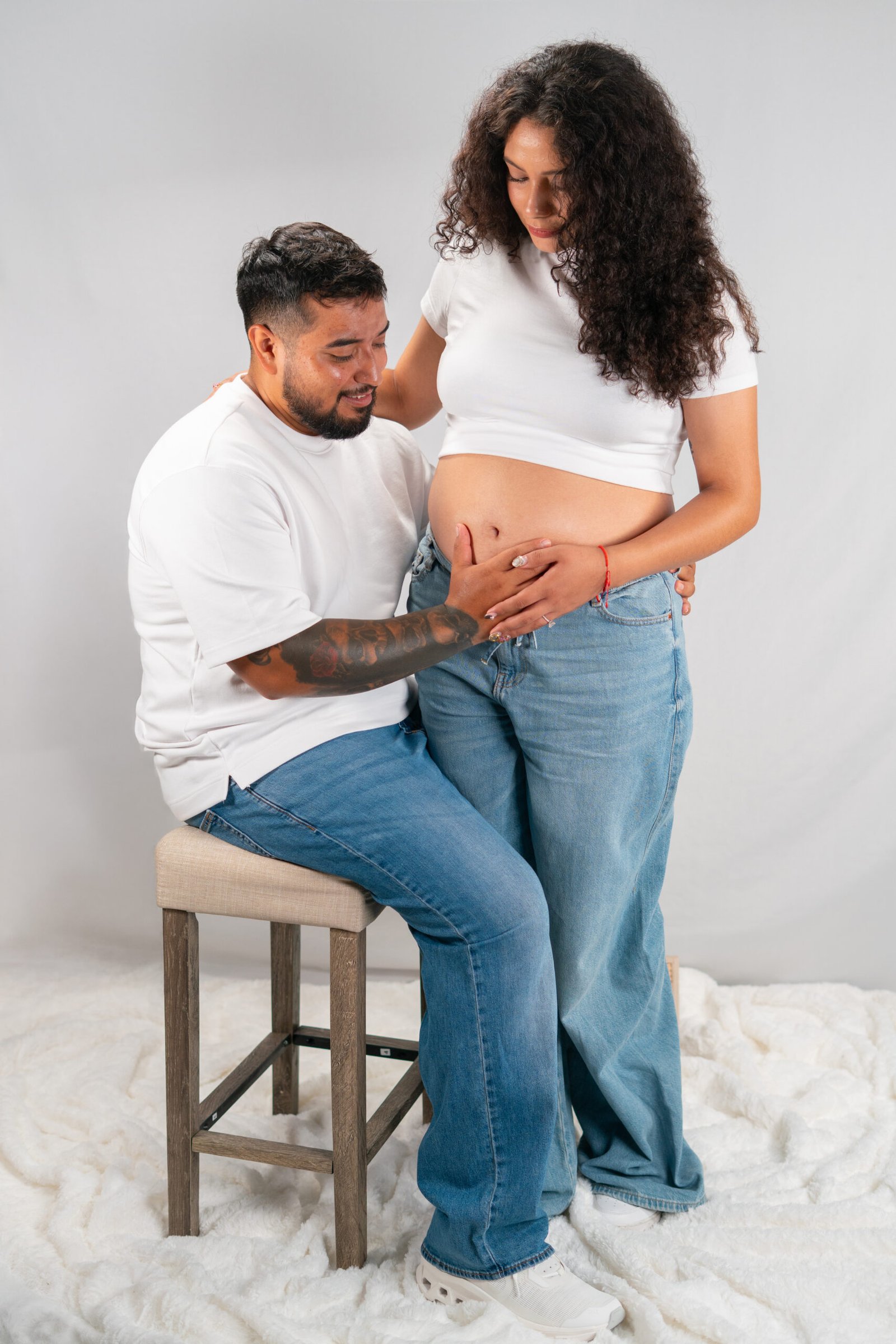 Pregnant woman standing beside her seated partner in a white studio portrait.