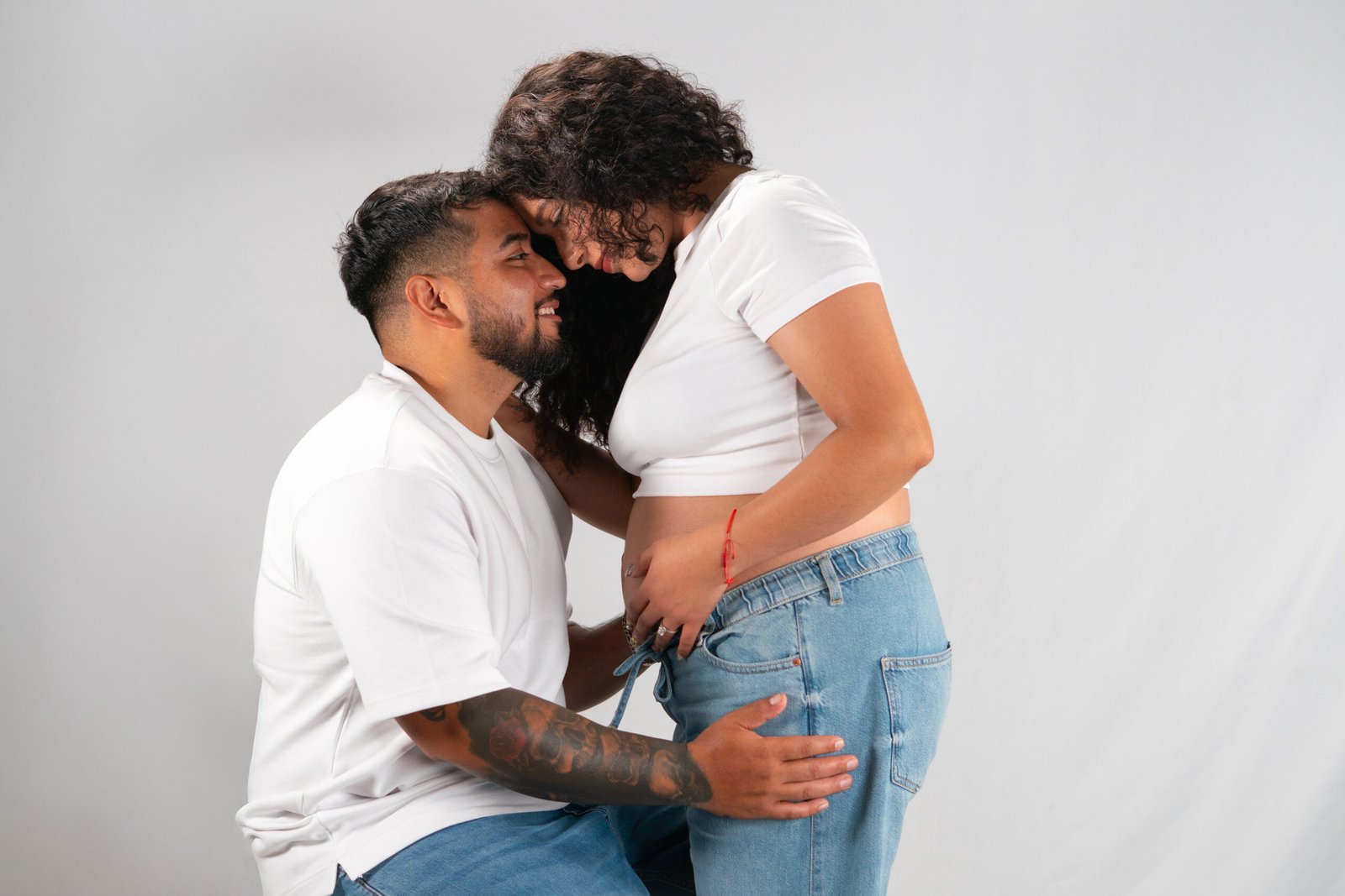 Expecting couple in white tops and jeans embracing against a white studio background.
