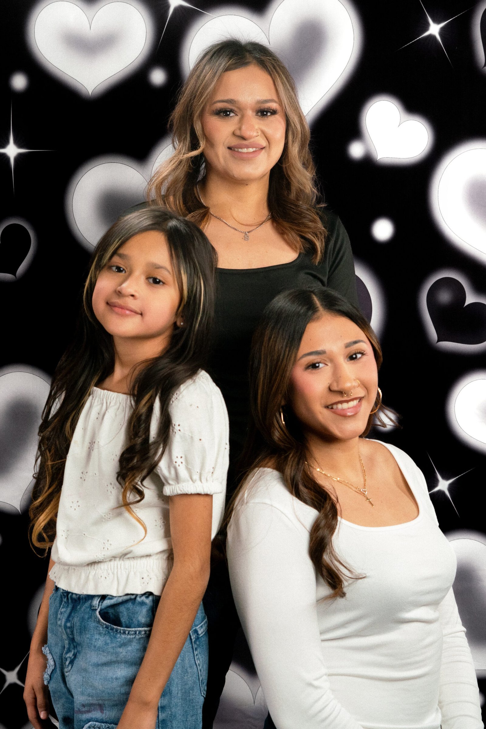 Three women pose in white and black tops against a glowing heart backdrop.