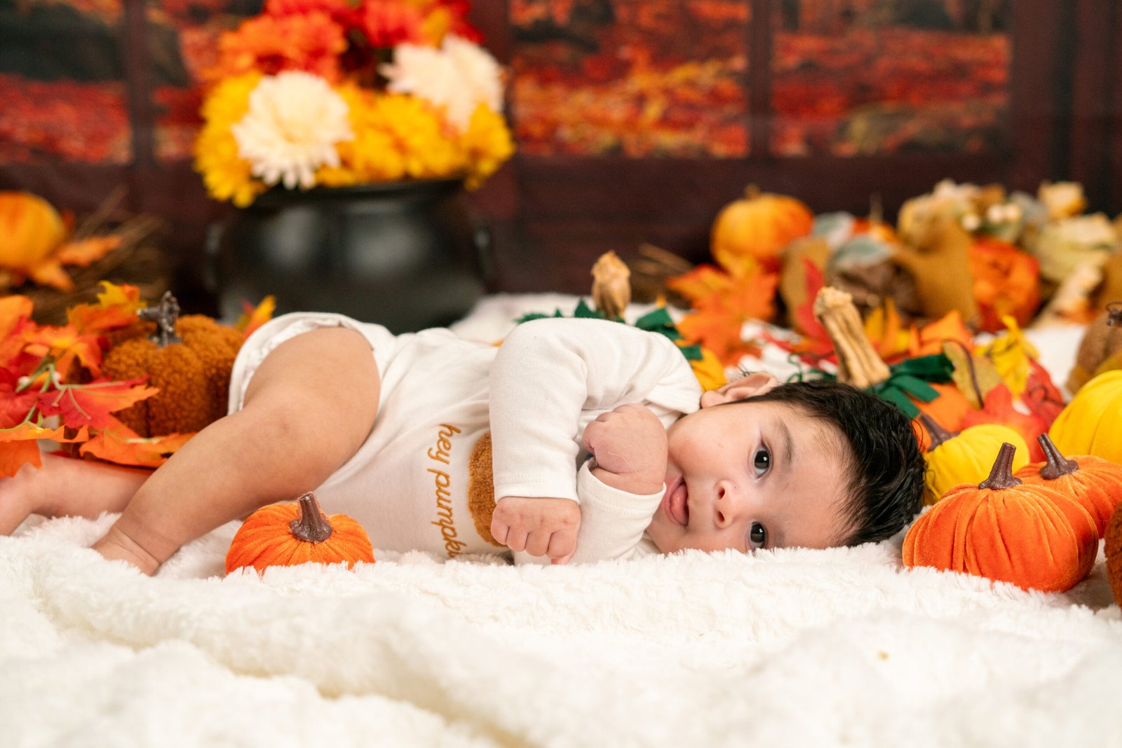 Baby in a white outfit lying among pumpkins and autumn flowers.