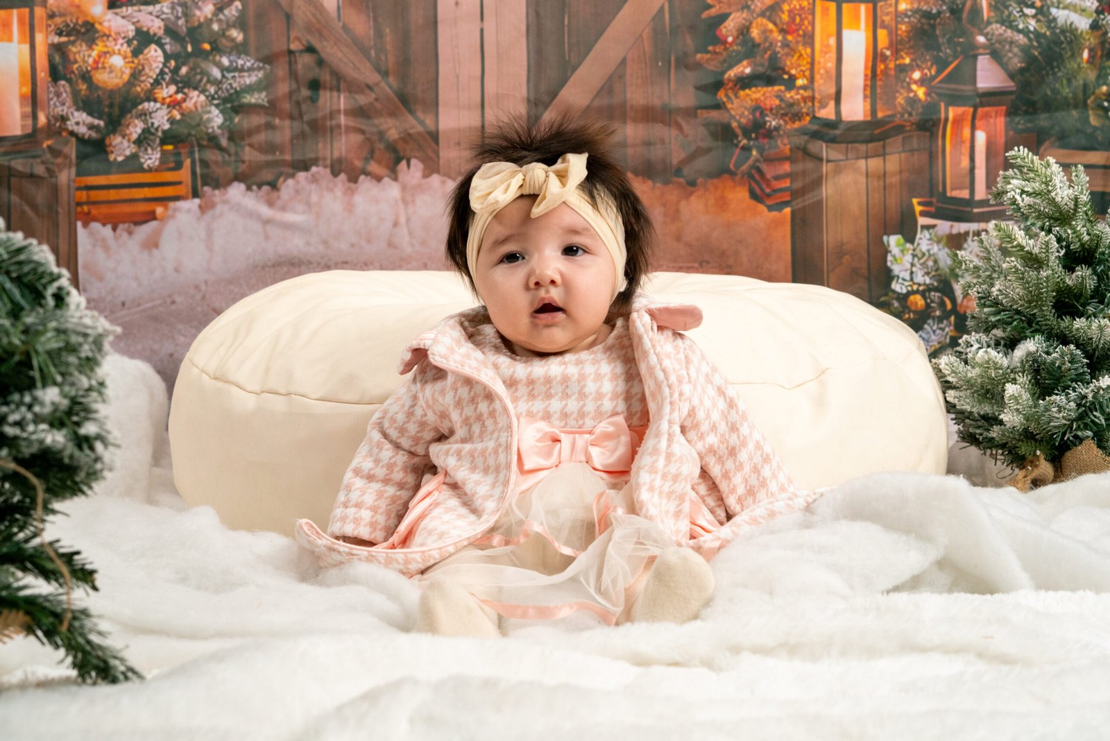 Baby in a pink outfit and bow seated on a white sofa in a snowy holiday set.